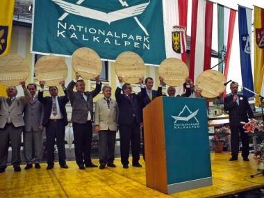 Politicians and municipal representatives stand on stage and hold up a wooden disk with the Kalkalpen National Park logo