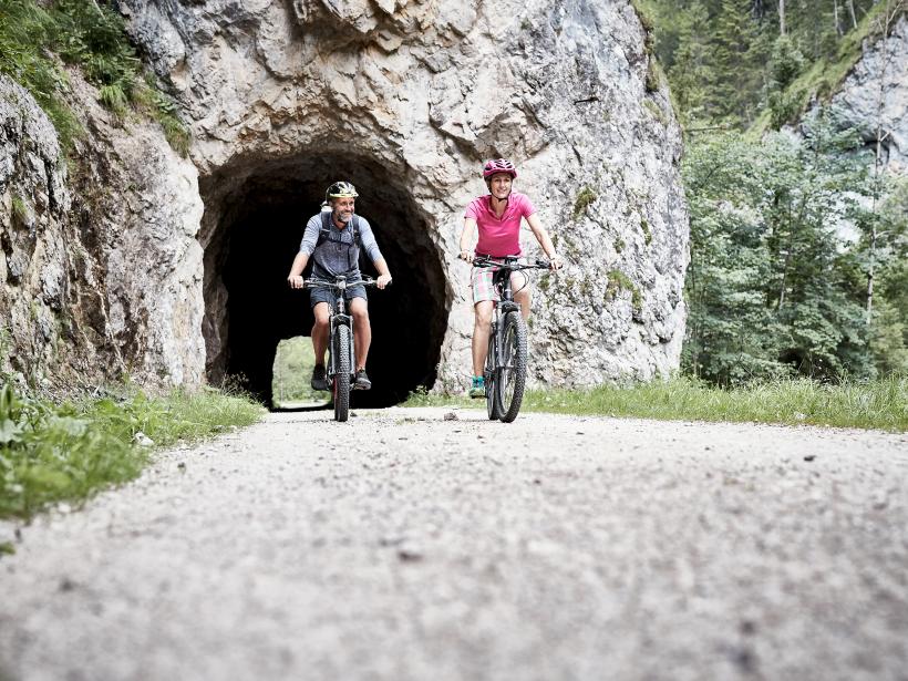 A woman and a man ride out of a rock tunnel on their mountain bikes