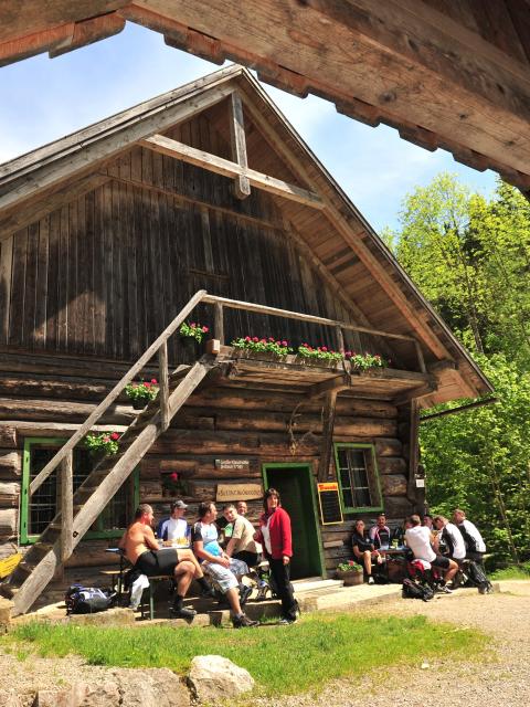 In front of an old log wooden building, people sit on benches and rest