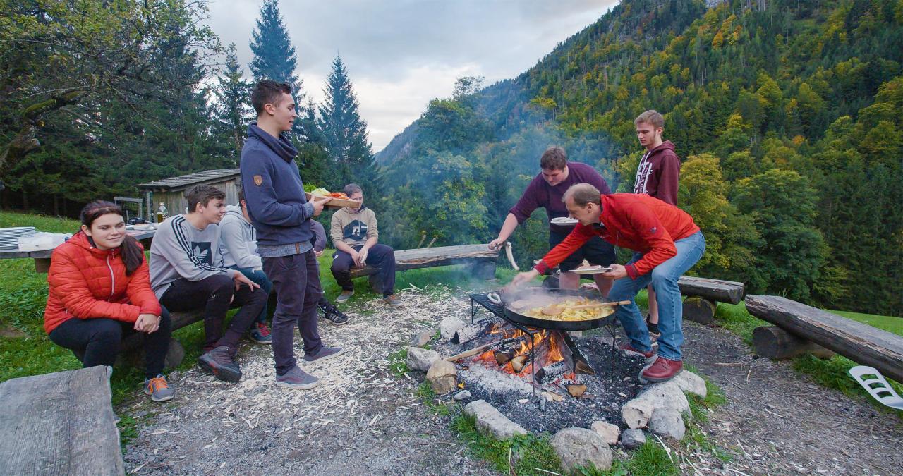 Young people sit around a campfire on which food is being cooked in a pan