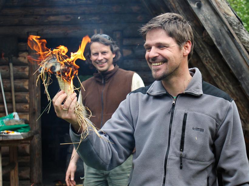 Two men light a fire in a piece of birch bark filled with grass