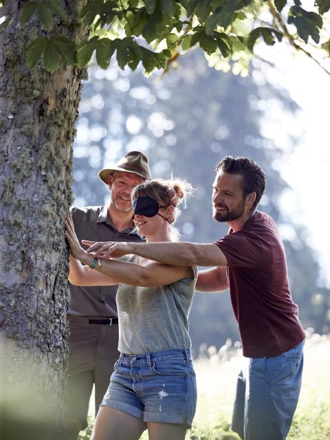 A blindfolded woman feels a tree trunk, a ranger and a man help with orientation