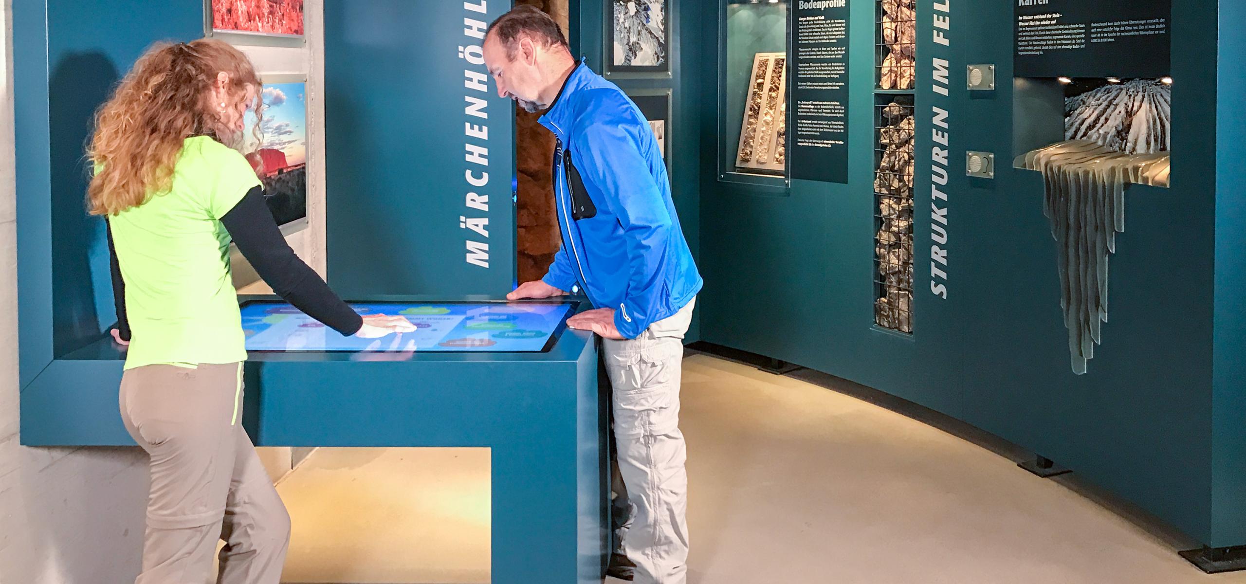 A woman and a man stand at a desk with a flatscreen in the Fascination of Rock exhibition
