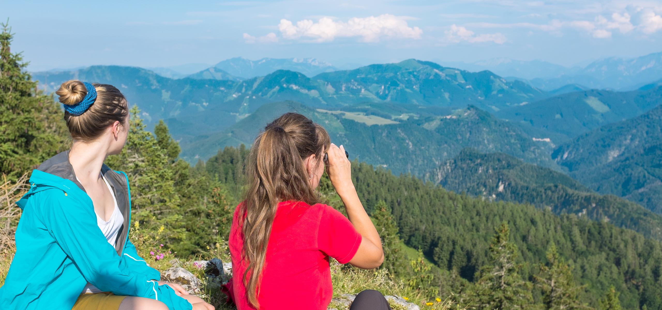 Two women sit on a mountain top and look over the mountain panorama