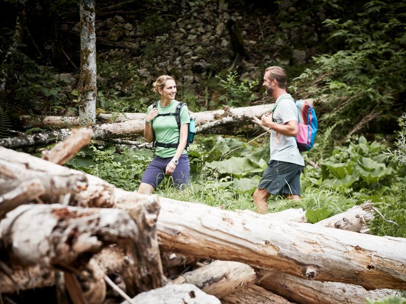 A woman and a man walk through a forest with fallen tree trunks